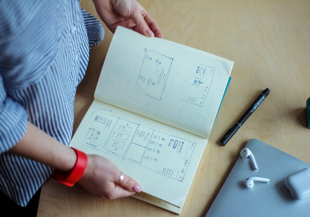 Person holding open software design sketchbook with pen, airpods and airpods case, and laptop on desk